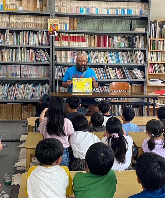 大勢の子どもたちのまえで紙芝居を披露する山本さんの写真。図書館かどこかでしょうか、背景にわ本がたくさん並んでいます。大勢の子どもたちの後ろ姿が映っています。まえのめりで聞いている子もいて、表情はみえませんがきっと笑顔で鑑賞しているのでしょう