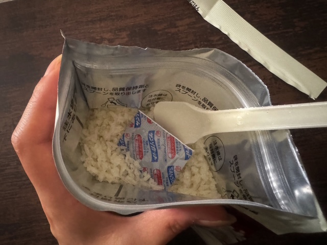 フクロをあけて品質保持剤とプラスチックのスプーンを取り出している様子を写した写真。白っぽいいろのプラスチックのスプーンと、コメの奥の方にあったしかくい品質保持剤をとりだします