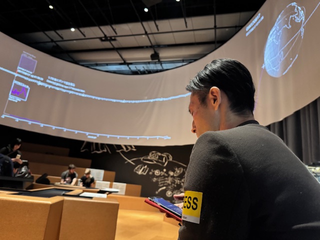 視覚障害当事者の男性が未来館の映像コンテンツをアシストアプリプラスで体験している様子を写した写真。男性の頭上には360度ぐるっと表示された大きなモニターがうつっています