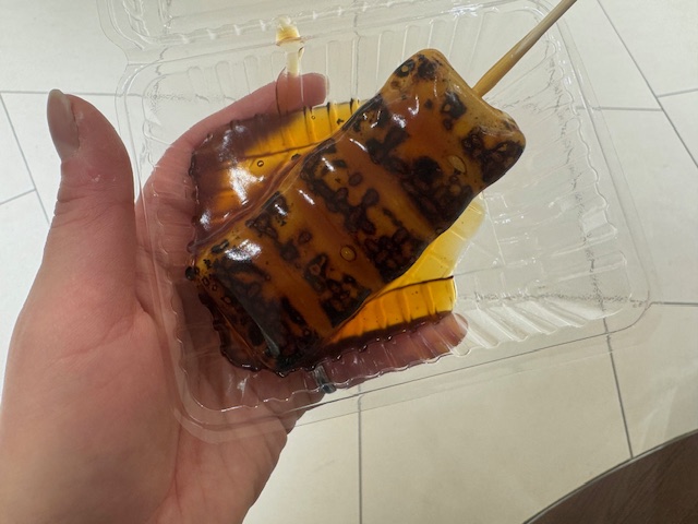 手でもった透明のプラスチックケースにいれられた1本のみたらし団子の写真。しっかりこげめがついたおだんごに、みたらしがたっぷりついています
