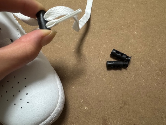 白いスニーカーのくつひもに、くろいストッパーをつけている様子を写した写真。スニーカーについたストッパー以外に、くろいストッパーふたつも映り込んでいます