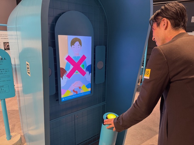 しらとりさんが、サトウの達人を体験している様子をうしろからうつした写真。この写真では、前方にあるモニターに、まるで病院の待合室のようなイラストが映しだされています。カルテをもった人が呼ぶ名前のなかから、サトウさんに聞こえたときに手前にある丸いボタンを押すというシンプルな設計。ちなみにボタンを押すと画面上の自分が、「ハイ」といわんばかりに片手を挙手します。写真ではミスしているので画面におおきくバツマークがついており、しらとりさんもあまりのむずかしさにくちもとがわらっています