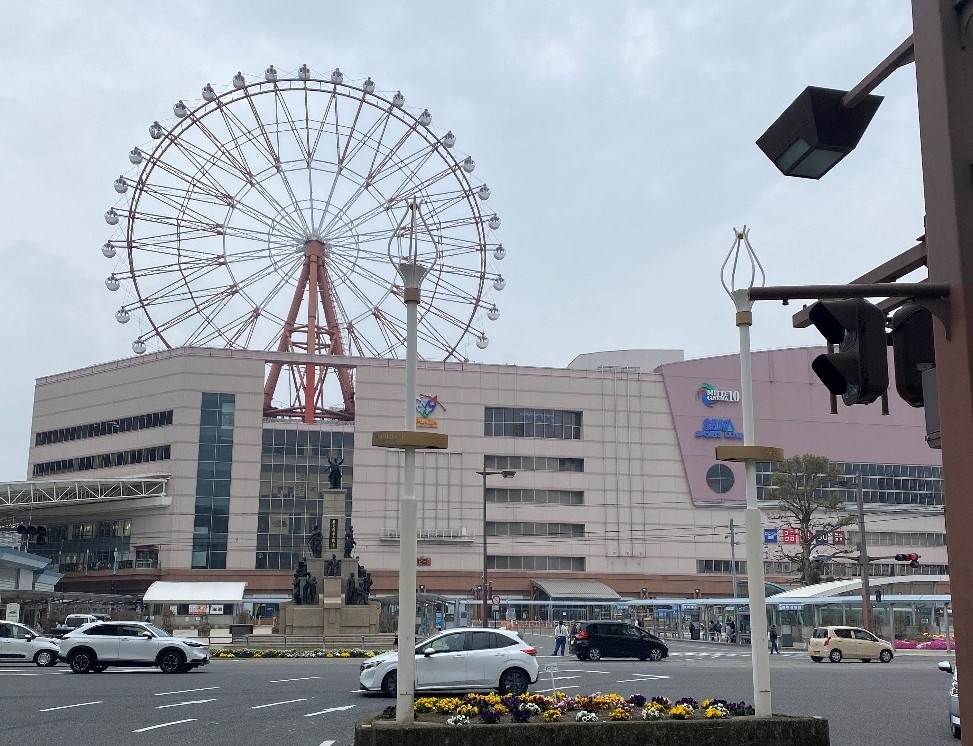 観覧車のある街並みが写っています。画像の中央には大きな観覧車がそびえ立っており、その手前には近代的なビルが建っています。ビルの壁面には、青とピンク色のロゴのようなものが確認できます。ビルの前には、車が行き交う道路が走っています。