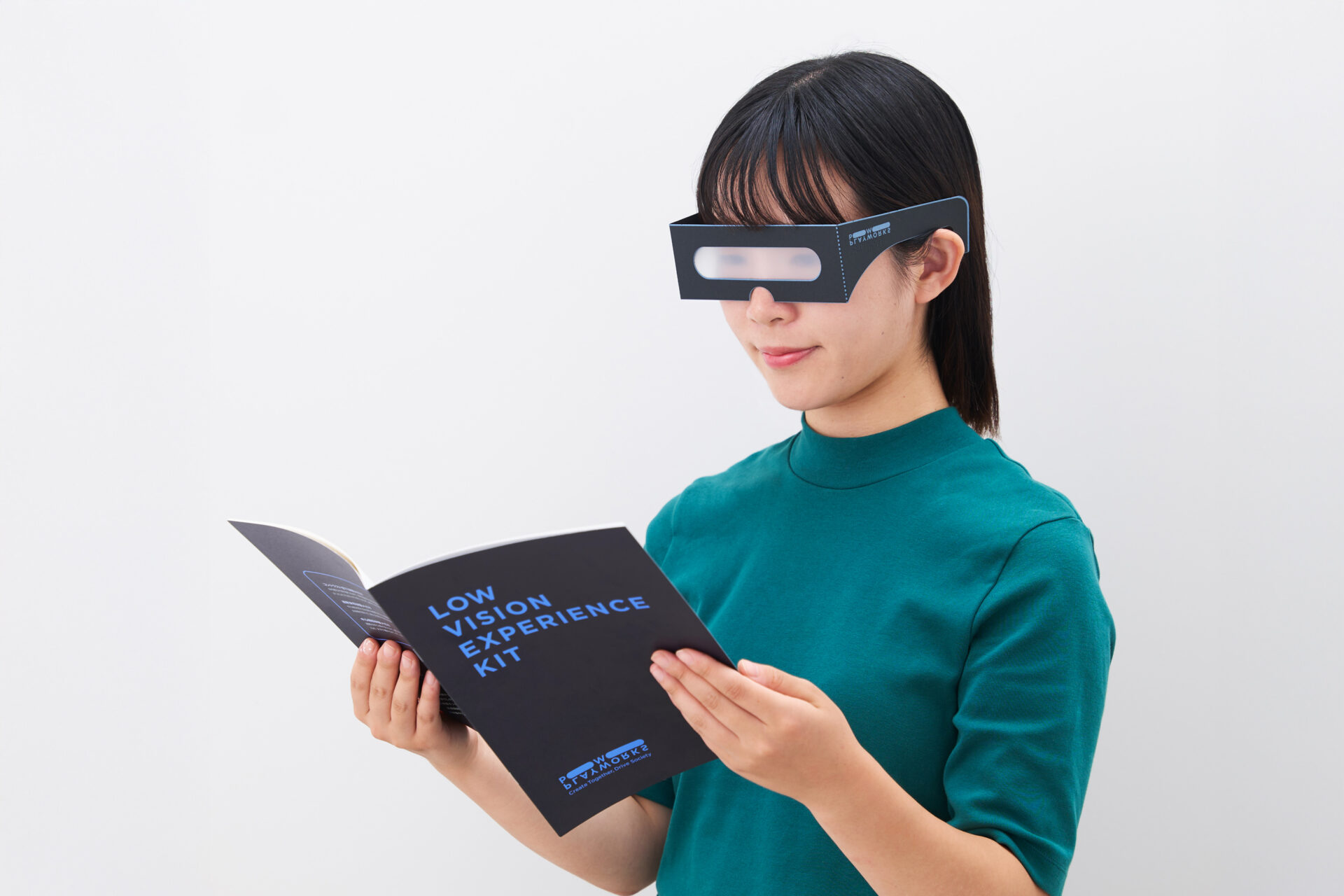 ロービジョン体験キットをつけて本を読んでいる女性の写真。ロービジョン体験キットわメガネがたで、しっかりした紙でできています