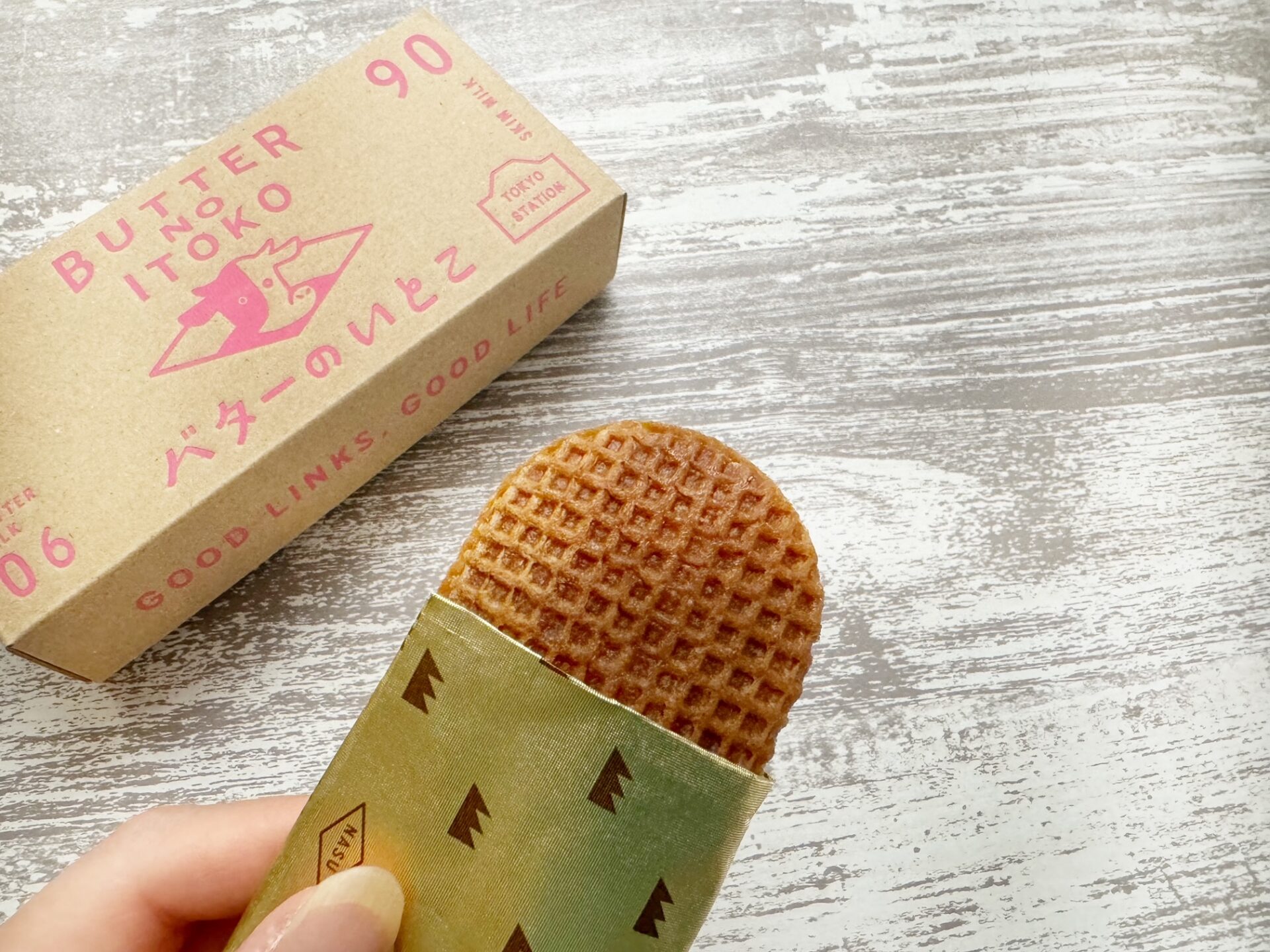 バターのいとこいちごチョコあじのパッケージを手でもっている様子と、そとハコの写真。紙製の箱にはピンクいろでバターのいとこという文字やイラストがかかれています。ハコを開けると3枚のバターのいとこが1枚ずつ紙につつまれ、さらに袋に入った状態でパッケージされています