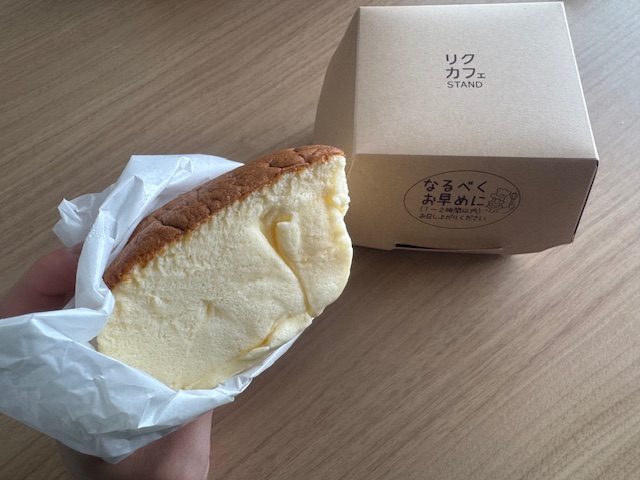 空港の休憩スペースのテーブルで、ハンバーガーみたいな紙に包まれたよんぶんのいちサイズの焼きたてチーズケーキをてでもった写真。背景にわハンバーガーのような紙製の箱もうつっています