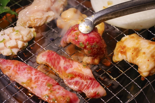 焼肉やさんのような場所で肉を焼いているてもとのイメージ写真。トングで肉をひっくりかえそうとしている様子がうつっています
