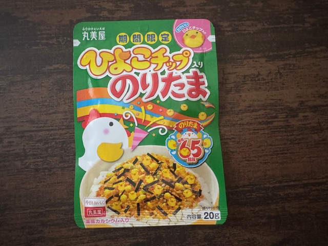 ひよこチップいりのりたまのパッケージ写真。グリーンベースのジッパーつきのふくろに、期間限定ひよこチップいりのりたまと文字が書かれています。ごはんにたっぷりかけられたのりたまの写真と、のりたま65周年のロゴ、クラッカーをパーンとした、横をむいたにわとりのイラストも描かれています。まるで65周年をおいわいしているかのようなにぎやかなパッケージです