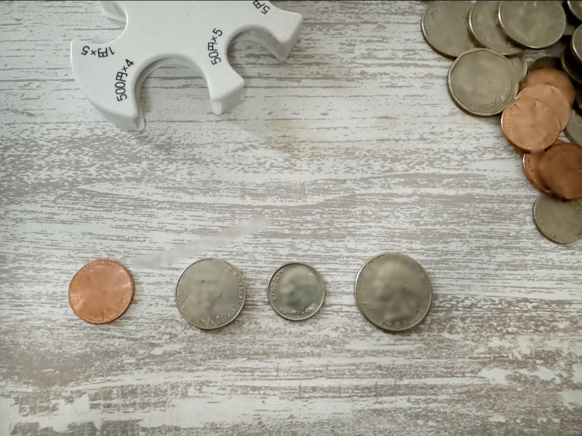 アメリカの硬貨、1セント、5セント、10セント、25セントを並べた写真。日本の硬貨とは大きさやあつさなどが異なります。背景にはしろいコインホルダーと、前回アメリカをおとずれたさいに使いきれなかったアメリカ硬貨の山がうつりこんでいます