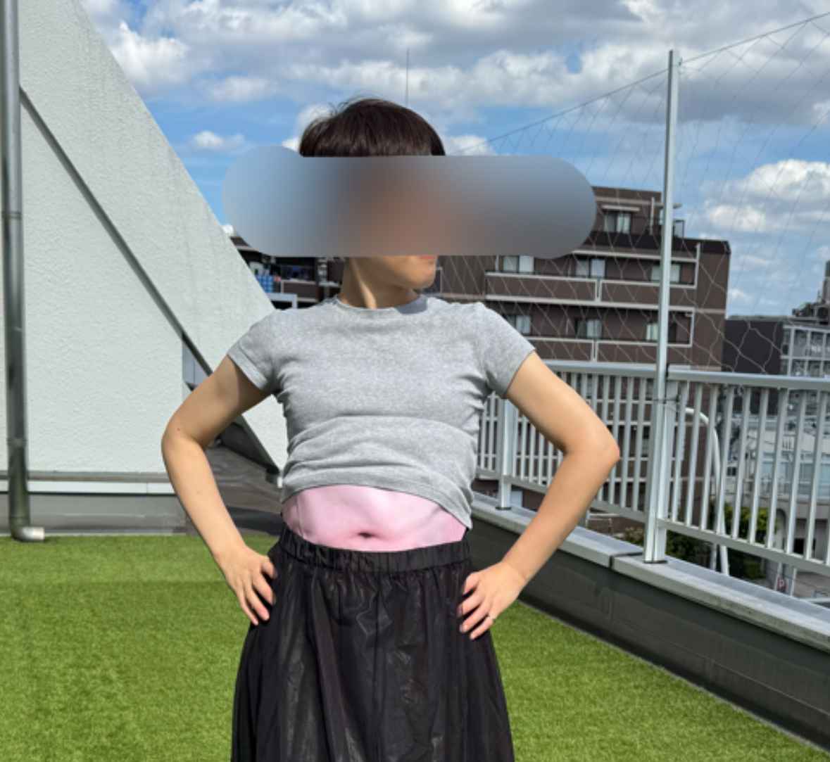 ウエストポーチをつけた女性のおなかの部分をクローズアップした写真。女性はピンクのウエストポーチに赤いパスポートと、黒い財布を入れています。グレーのTシャツを着ており、黒っぽいスカートのようなものを身に着けています。背景には、窓や緑色の芝生の一部が見えます