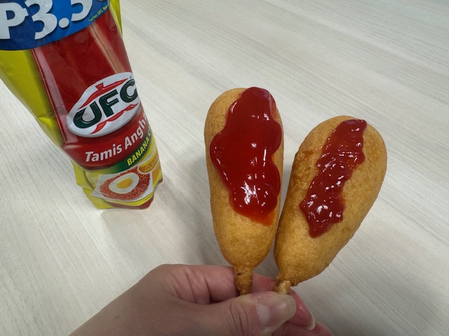 2本のアメリカンドッグをてでもっている写真。1本わバナナケチャップをかけており、もういっぽんわトマトケチャップをかけました。バナナケチャップのほうがじゃっかんみずっぽくみえるものの、写真でみるといろもほぼ同じで、その違いがほとんどわかりません。いってしまえば、それくらいみためわそっくりということでしょう ハイゴにわふくろにハイッタリフィルのバナナケチャップの黄色いパッケージがうつりこんでいます