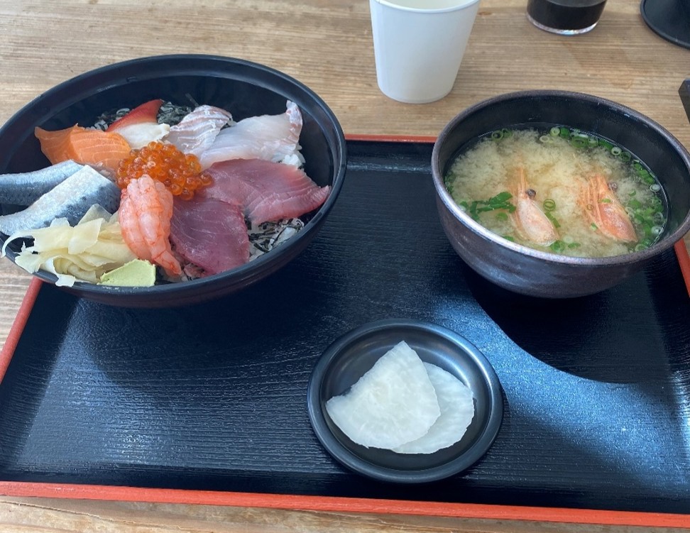 彩り豊かな海鮮丼と、エビの入ったお味噌汁、そしてお漬物が並べられた、穏やかなひとときを感じさせる食事風景です。最も目を引くのは、黒いどんぶりに盛り付けられた海鮮丼です。中央には輝く宝石のようなイクラが添えられ、その周りにはマグロ、サーモン、白身魚、エビがまるで海からの贈り物のように美しく並んでいます。どんぶりのそばには、生姜の甘酢漬けとわさびが添えられています。海鮮丼の右隣には、刻みネギと有頭(ゆうとう)エビが入ったお味噌汁が、温かみのある黒い椀に盛れられています。これらは木目調のテーブルの上に配置されており、全体として落ち着いた和の雰囲気が漂っています。