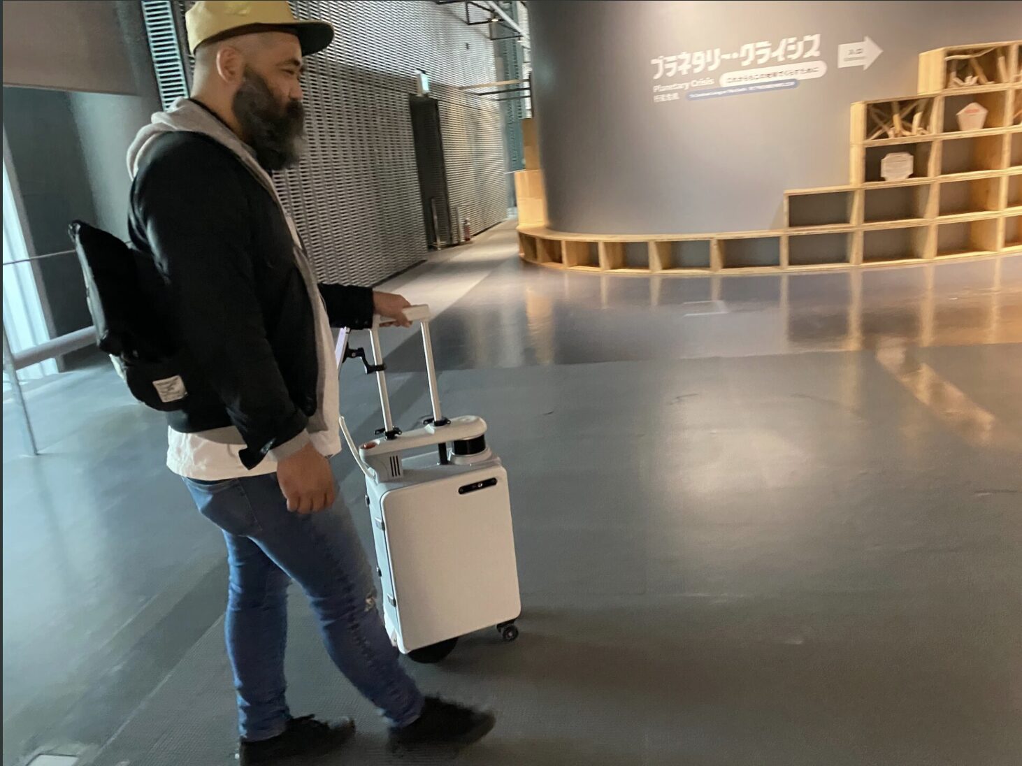 全盲の当事者の山本さんが、未来館のなかでエーアイスーツケースに誘導してもらっている過去の写真。あたまにわキャップをかぶっています