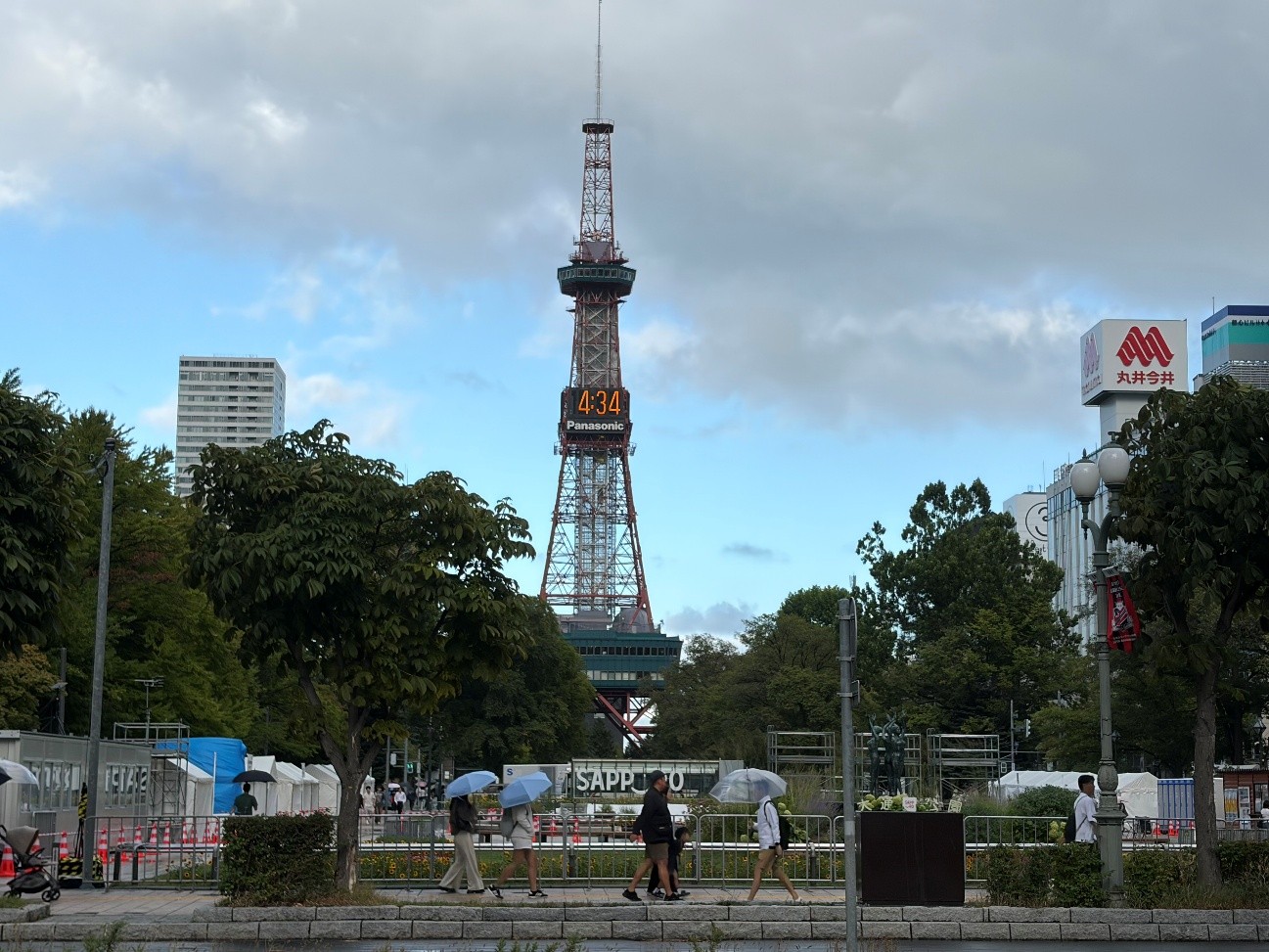 札幌のテレビ塔がそびえ立つ大通公園の様子です。塔の中央付近にはパナソニックのロゴが表示されたデジタル時計があり、手前には歩行者や仮設の柵が見えます。撮影時のデジタル時計は「4時34分」と表示されています。画面右側には、白い看板に赤い文字で「丸井今井」と書かれた建物が見えます。また、中央下部には「サッポロ」と書かれたロゴパネルが設置されています。テレビ塔の周囲には緑豊かな木々が茂っています。空は雲に覆われていますが、所々に青い空がのぞいています。