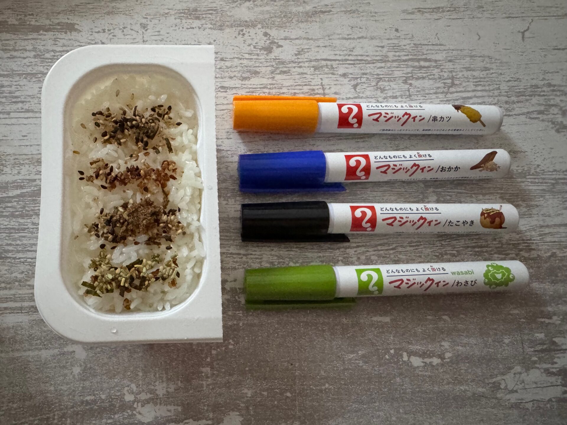 インスタントのレンチンごはんのうえに、うえからオレンジ、あお、くろ、みどりのふりかけを直線になるように、まるでスケッチブックによこ線を一本引くようにふりかけた写真。なお、レンチンごはんのようきの右にわうえからオレンジ、あお、くろ、みどりのふりかけ本体が並んでいます