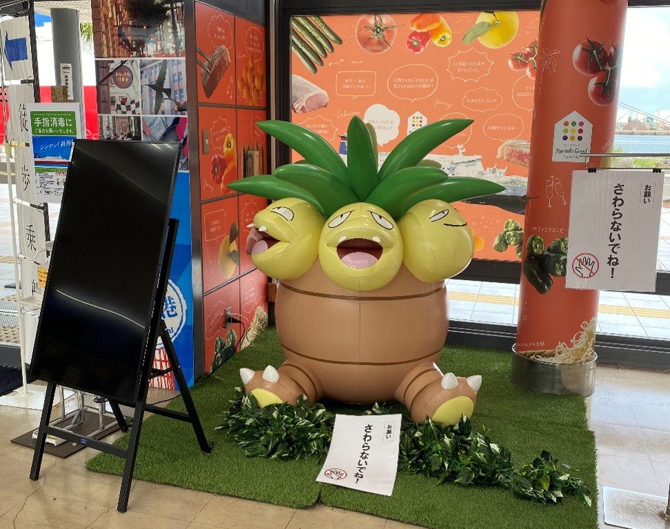ポケモンであるナッシーの大きなフィギュアが飾られている様子です。中央に鎮座するナッシーは、ヤシの葉のような形状の髪型と、頭部が3つあり、それぞれ異なる表情を浮かべています。その下には樽のような形状の胴体に太い足が生えています。フィギュアは人工芝のような緑色のマットの上に置かれています。