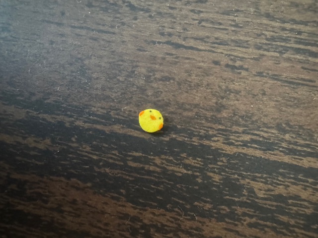 ひよこチップひとつをうつした写真。ちょっけいすうミリ程度のきいろくてちいさなまるいチップに、オレンジでくちばしとりょうて、くろくてつぶらなひとみがえがかれています