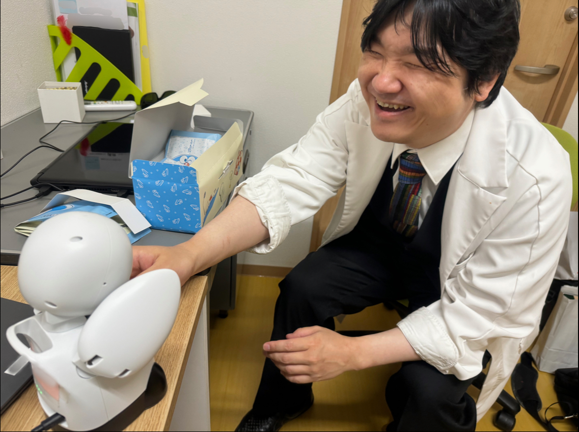 インタビューをお願いした福場先生が勤務する病院の診察室で、オリヒメごしに先生と会話している様子を移した写真。オリヒメと先生が握手しています。先生もものすごくいい笑顔です