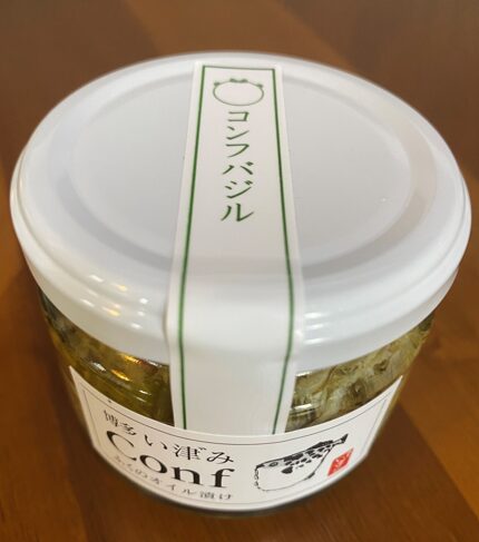 ふぐのオイルづけ、コンフバジルふうみの瓶の写真。瓶は白い蓋で密封されており、なかにはオイル漬けされたふぐの身が見えます。瓶には白いラベルが貼られており、縦長のラベルには「コンフバジル」と書かれています。ラベルの上部にはふぐのイラストが描かれています。その下の正面には白いラベルが貼られており、上部に「ハカタいづみと日本語が書かれ、そのしたに大きな文字で「Conf」と印字されています。さらに下には「ふくのオイル漬け」と書かれ、右下にはふぐのイラストと赤い印鑑がデザインされています