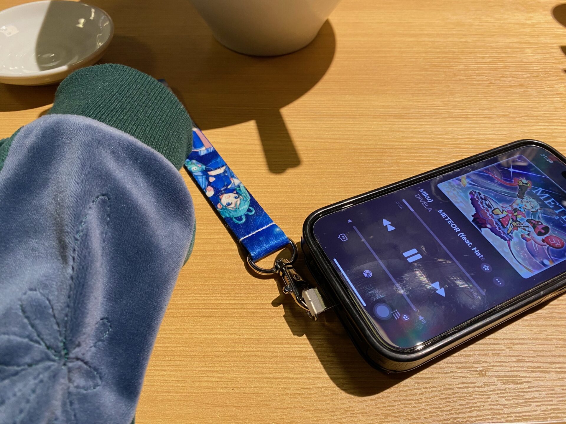 初音ミクのベロアのツアージャケットと、携帯のストラップも初音ミク、スマホの画面は、さっきまで聞いていた初音ミクのミーティアという曲のジャケット画像。奥にはラーメンのうつわも映り込んでいます