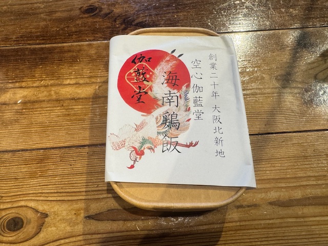くうしんがらんどうの駅弁、かいなんけいはんのパッケージ写真。かどがまるいちょうほうけいのちゃいろい容器に、「創業20年、大阪きたしんち、くうしんがらんどう、かいなんけいはん」とかかれた紙でくるっとおおわれており、中身が見えません