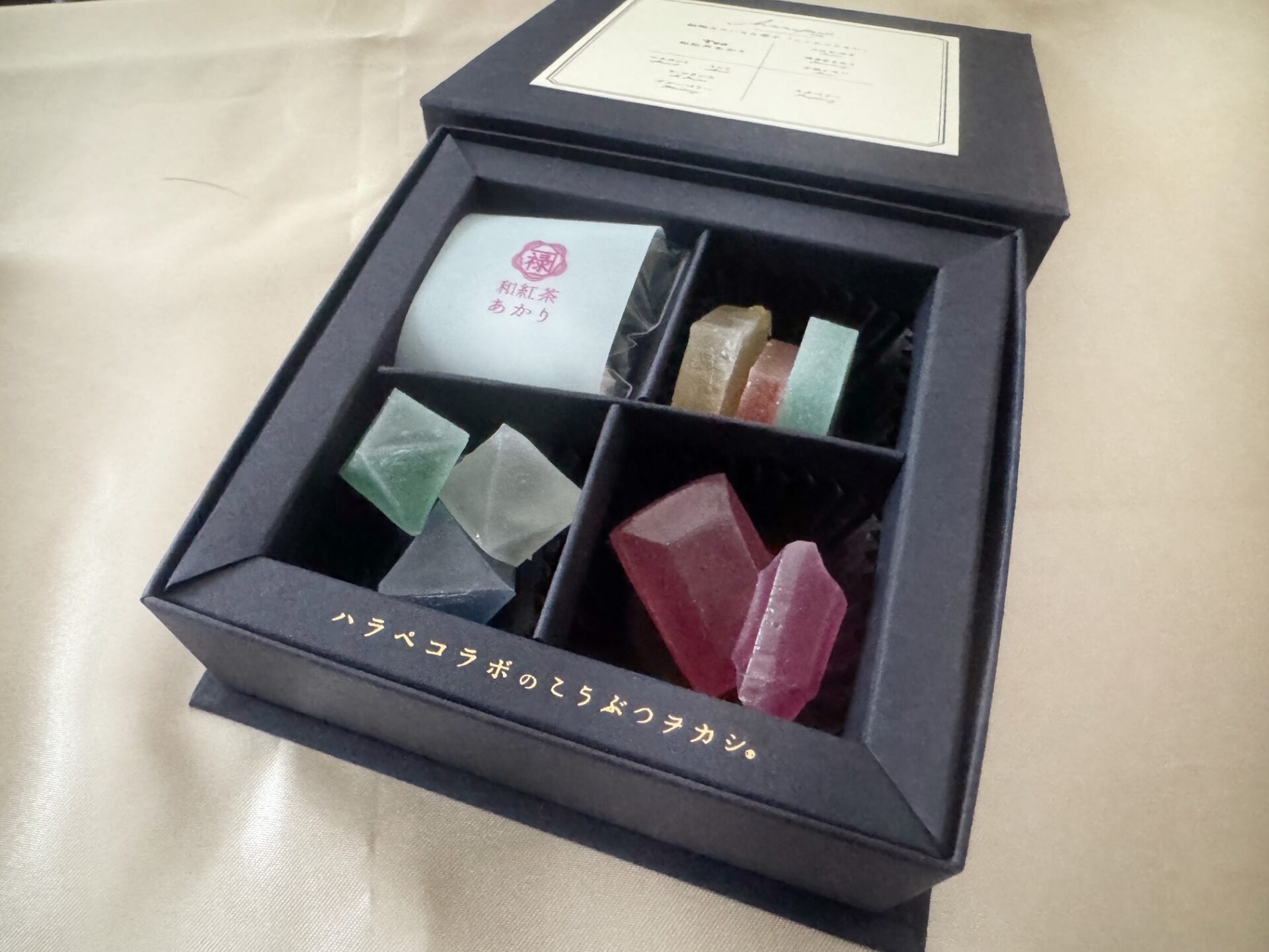 こだわりのこうぶつヲカシと和紅茶セットの紙製のネイビーのハコのふたをあけた様子を写した写真。なかわよっつにくぎられており、ひだりうえに3グラムいりのティーバッグがハイッタ紅茶のパッケージ、みぎうえが、おおいたかぼす、ハカタあまおう、うきレモンのこうぶつヲカシがひとつずつ、右下が宝石カットのあかいラズベリーのこうぶつヲカシがふたつ、ひだりしたにわマスカット、ミント、ラフランス、ブルーベリーのセイハチメンタイのりったいてきなこうぶつヲカシがひとつずつハイっています