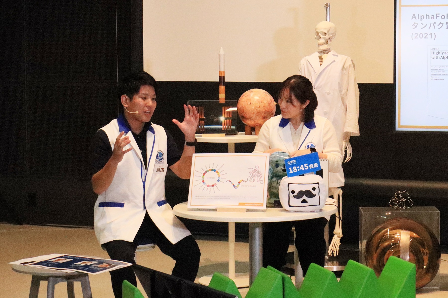 昨年の配信の様子を写した写真。男女ふたりの科学コミュニケーターがいすにすわり、テーブルを囲みながら解説をしています。テーブルのうえには解説用のパネル、ニコニコテレビちゃんのぬいぐるみには黒ひげがついています。背後にいる人体骨格もけいは白衣をまとっています