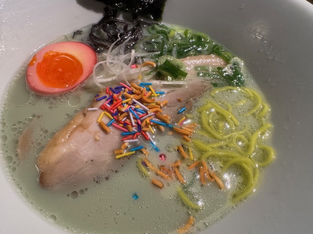ラーメンにトッピングを載せた写真。緑のスープ、ピンクのたまご、カラフルなシュガースプレーとベビースター。カラーリングがラーメンのアレではない