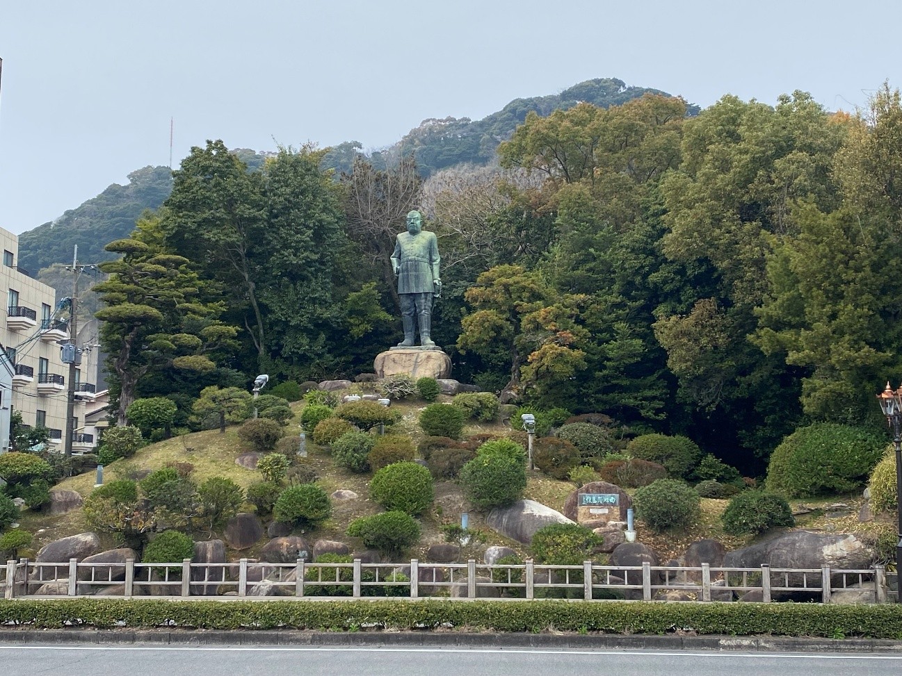 丘の麓に立つ銅像と手入れされた庭園の様子が写っています。庭園には、丸く刈り込まれた植栽が点在し、石も配置されています。庭園の手前には白い柵があります。銅像は、軍服のようなものを着ており、堂々とした立ち姿で、全体的に静かで落ち着いた雰囲気が漂っています。