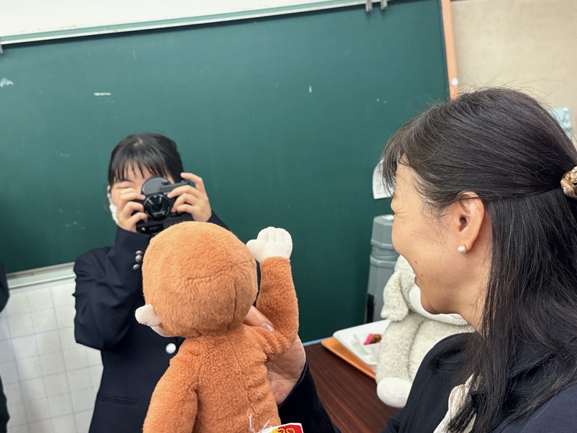 網膜投影カメラキットを手にした女の子の前で、先生がサルのぬいぐるみを持って立っている様子をうつした写真。女の子はカメラごしにぬいぐるみをみています