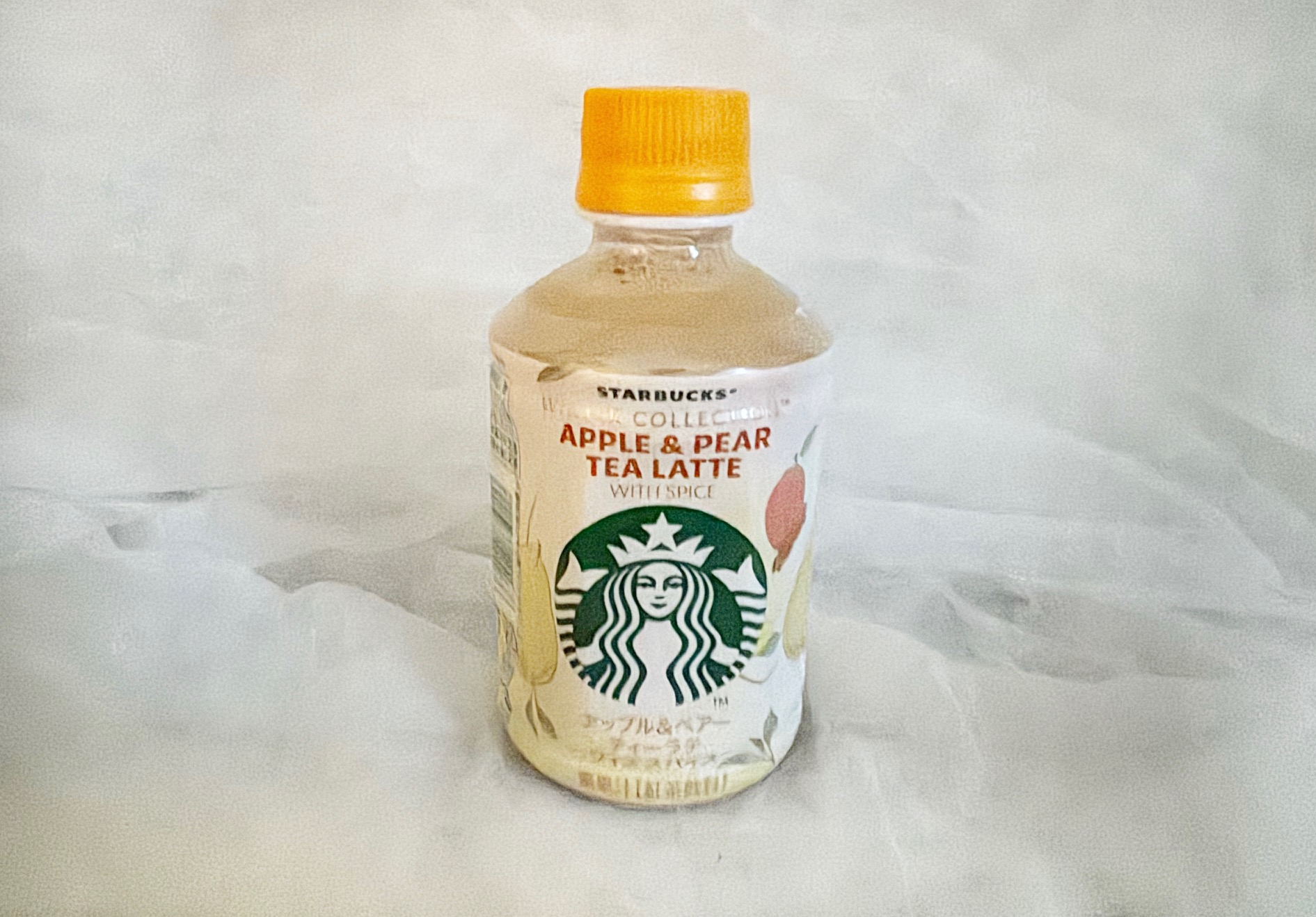 スターバックスウインターコレクションアップル&ペアーティーラテ ウィズスパイスの商品写真。白っぽいラベルにあかいりんごやみどりいろのナシのイラストがえがかれており、中央にはスターバックスのロゴがあしらわれています。ペットボトルのフタはオレンジです