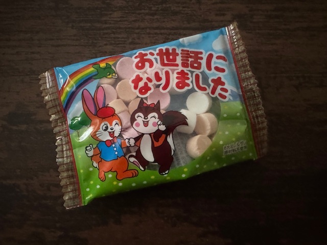 グッピーラムネの個包装の写真。あかいもじで「おせわになりました」とかいてあり、レトロなうさぎとリスが、そうげんでなかよくてをつないでいるイラストがえがかれています。ハイゴにわ虹がかかっており、みどりいろのことりもとんでいる、にぎやかなイラストです。とうめいな部分からわカラフルなラムネがのぞいています