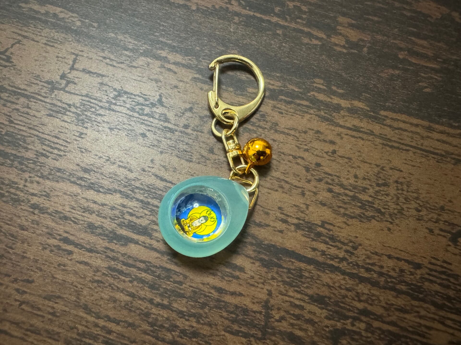 キーホルダーじょうのおまもり、しずくの写真。ひとしずくのしずくをかたどったみずいろのキーホルダーで、ゴールドのナスカンがついています。しずくにはレンズがついており、なかに御本尊のお姿がみえます