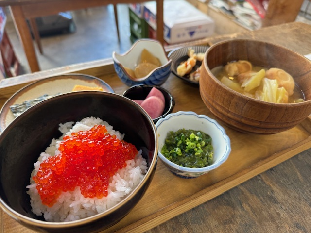 木製のおぼんにのった、すじこめしの写真。くろいおちゃわんにもられたハクマイと、きらきらかがやくすじこや、きのおわんによそったぐだくさんのしるもの、おつけもののコバチなどがならんでいます