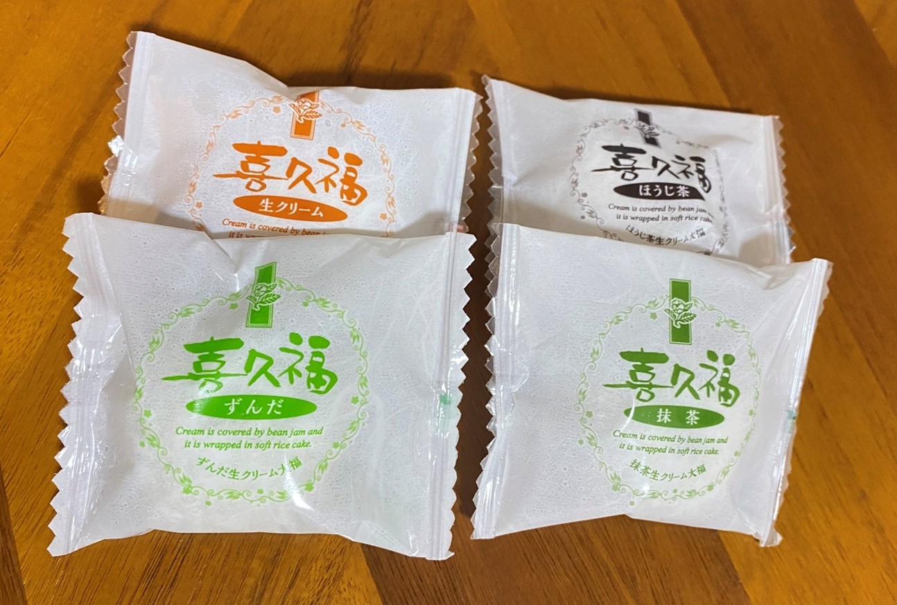 箱には和菓子が4つ、白い個包装に包まれて入っています。個包装にはそれぞれ喜久福(きくふく)という文字と、味を表す漢字が印字されています。左上の個包装にはオレンジ色の文字で生クリーム、左下の個包装には緑色の文字でずんだ、右上の個包装には黒色(くろいろ)の文字でほうじ茶、右下の個包装には緑色の文字で抹茶と記されています。