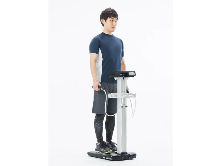 タニタの靴を履いたまま測定できる、プロフェッショナル仕様のたいそせいけいにのった男性の写真。両手に電極のついたバーをにぎっています