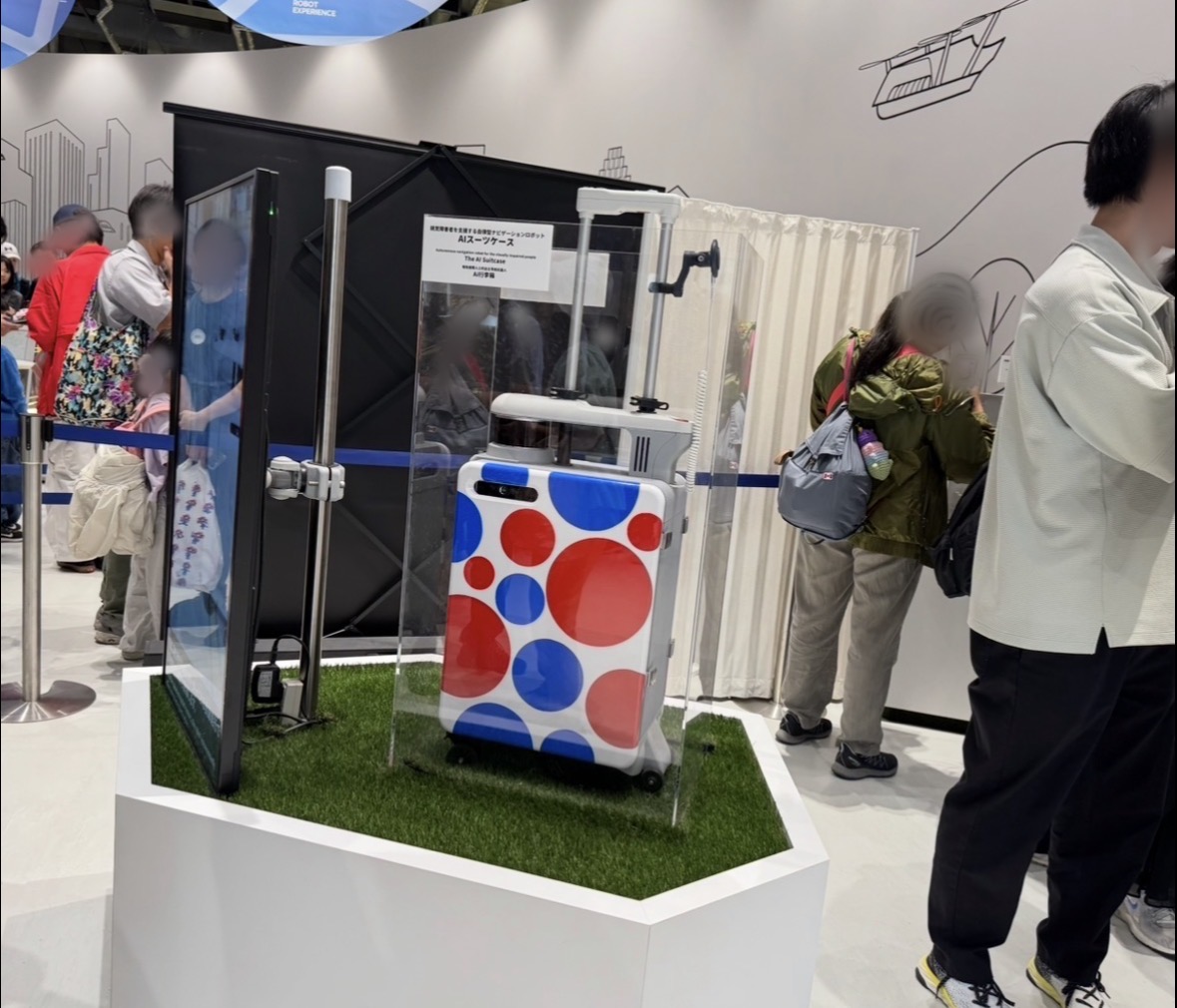 大阪・関西万博の会場、展示会場で撮影したエーアイスーツケースの写真。背後にわ、すうめいの人が写り込んでいます。エーアイスーツケースわ赤と青の大きなみずたまもようが特徴的で、グリーンの人工しばがしかれた、白い台のうえにおかれています