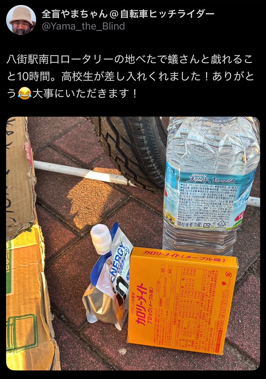 山本さんのXの投稿のスクリーンショット。「やちまたえきみなみぐちロータリーのじべたでアリさんと戯れること10じかん。高校生がさしいれしてくれました！ありがとう。大事にいただきます」とテキストがかかれており、じべたにおかれたカロリーメイト、ペットボトルのみず、ウィダーインゼリーがひとつずつと、ダンボール、ハクジョウ、タンデムのタイヤらしきものもうつりこんでいます