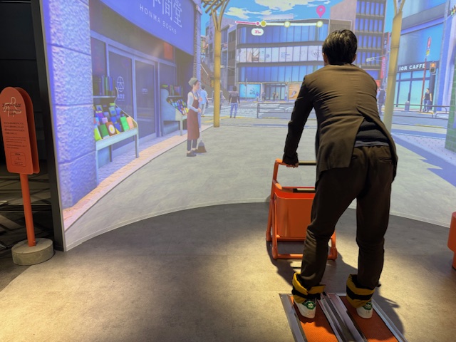 「スーパーへゴー!」を体験しているしらとりさんの後ろ姿の写真。手前に広がる巨大スクリーンには風景イラスト、手前にはカートがあり、体験する人は、あしにおもりをつけて老化を疑似体験しつつ、その場でカートを押しながら足踏みしてスーパーまで向かいます