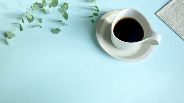 テーブルにのったコーヒーの写真。かんようしょくぶつのハッパと、えいじの新聞がうつりこんでいます