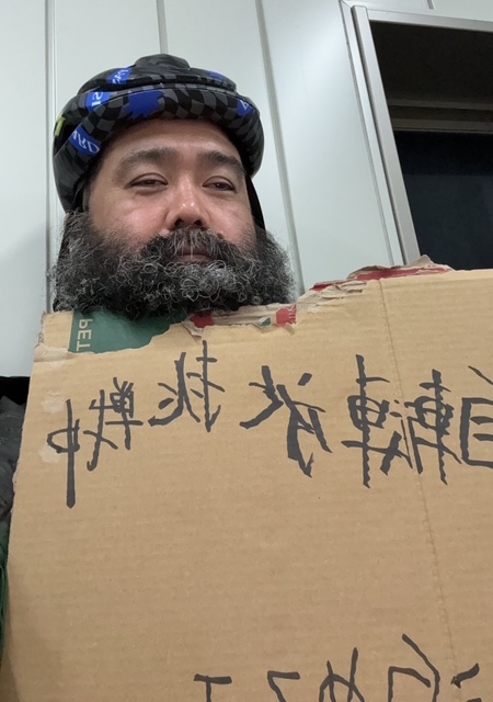 ダンボールに「自転車旅挑戦中」と手書きの文字で書かれたものを山本さんが持っている写真