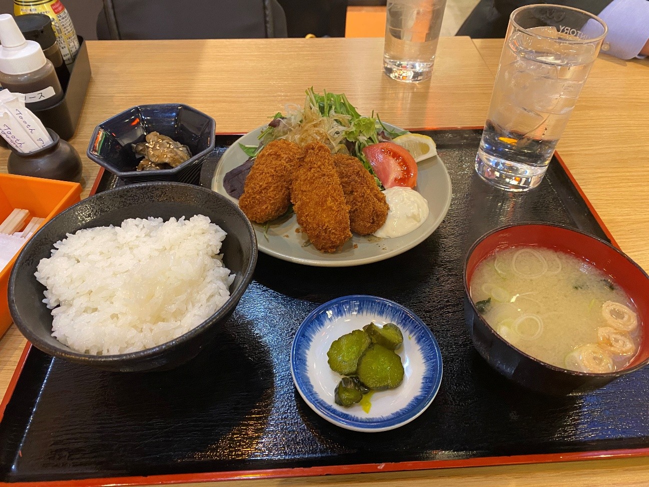 牡蠣定食です。お盆の上には、ご飯、揚げ物、お味噌汁、漬物が並んでいます。ご飯は白米で、ふっくらと炊きあげられています。牡蠣フライは衣がカリっとしており、食欲をそそるきつね色をしています。お味噌汁は具材にネギが入っており、温かい雰囲気が伝わってきます。漬物はきゅうりのピクルスで、彩りのアクセントになっています。全体的に、家庭的でほっとするような定食の画像です。