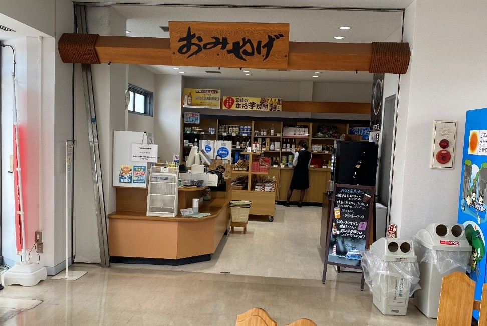 落ち着いた雰囲気の店内で、お土産品や本格焼酎などが販売されている様子です。入り口上部には木製の看板が掲げられており、右から左に向かって「おみやげ」と力強い筆文字で書かれています。その奥の棚の上部には「宮崎の本格焼酎」と記された黄色い横長の看板が設置されており、本格焼酎の銘柄や酒類が豊富に並んでいるのが見受けられます。