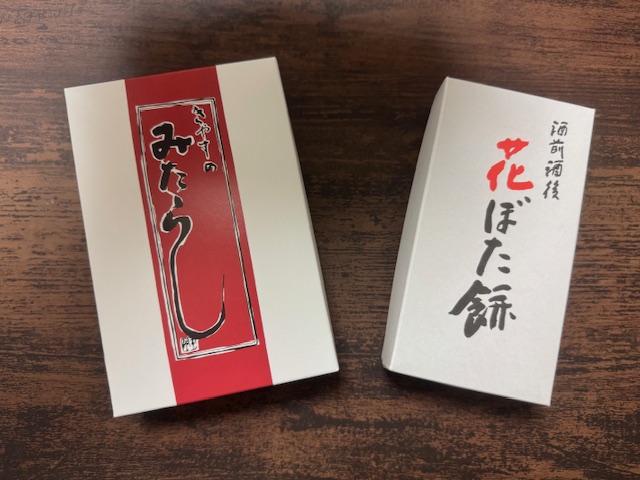 みたらし団子5本入りと、ハナぼたもち2個いりのパッケージ写真。どちらも紙製のハコにハイっています。両方とも白いハコで、ふでがきふうのもじで商品めいがかかれています