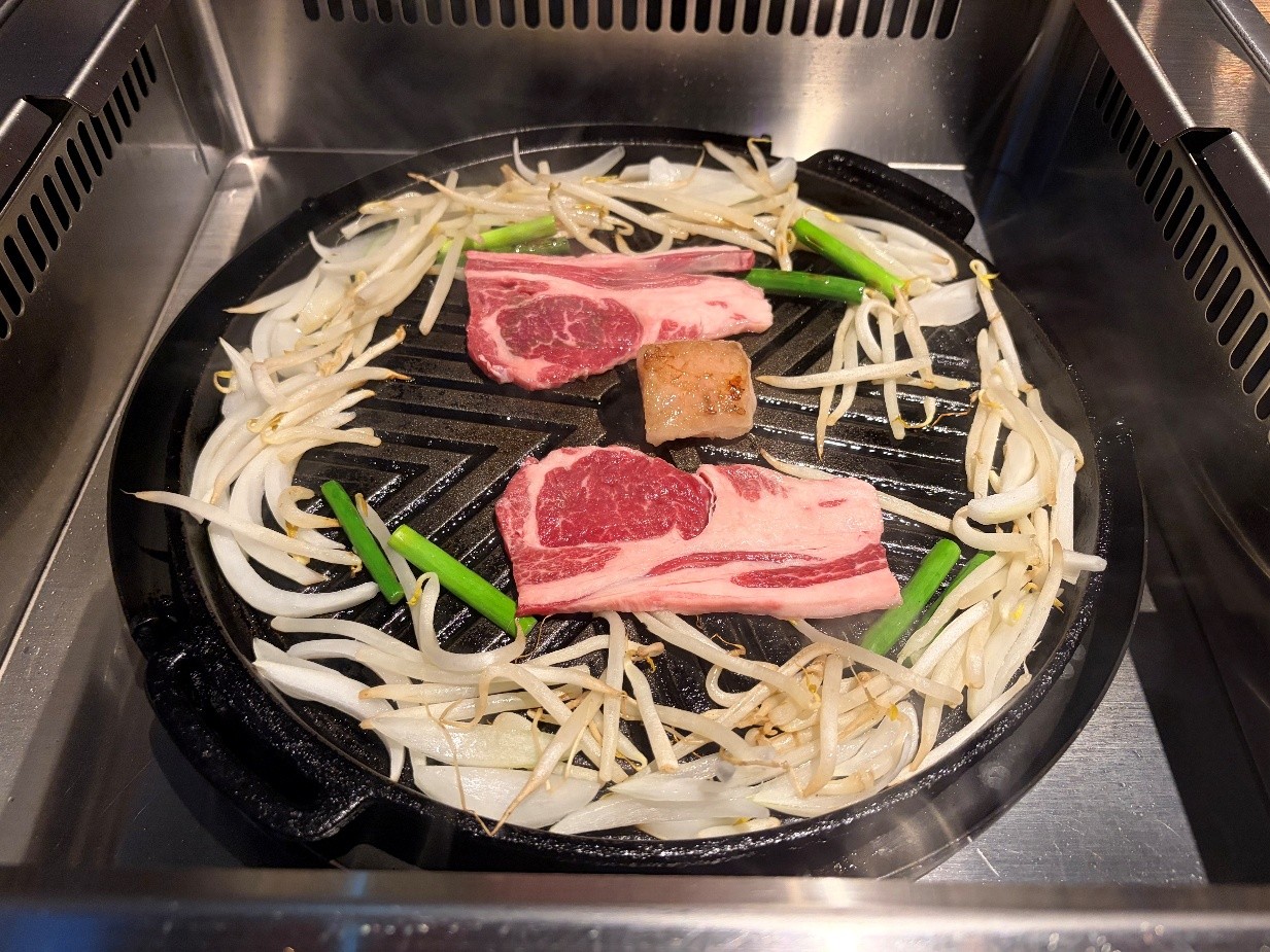 ジンギスカン鍋で羊肉と野菜を焼いている様子です。中央の鍋には肉と野菜が並べられ、調理が進んでいます。 中央には、赤身と脂身がはっきりとした二枚の羊肉が置かれています。その間には、焼くための脂の塊が一つ配置されています。羊肉の周囲を囲むように、もやしと玉ねぎ、そして緑色のねぎが鍋のフチに沿って敷き詰められています。 鍋は黒い鋳物製で、中央が盛り上がった独特の形状をしています。鍋の周囲は金属製のコンロ台になっており、調理中の熱気が伝わってくるような雰囲気です。