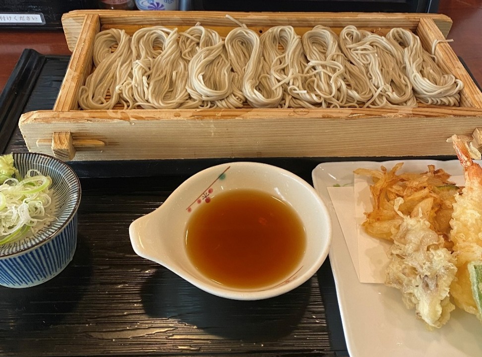 やや大きな器に盛られたへぎそばと天ぷら、つゆが並ぶ食卓の様子です。中央には、木製の長方形の器に一口サイズに丸められたそばが8つ並んでいます。その右側には白い皿に盛られた天ぷらが置かれ、左側には天つゆが入った小鉢(こばち)と、薬味のネギを乗せたそばつゆの小鉢(こばち)が配置されています。