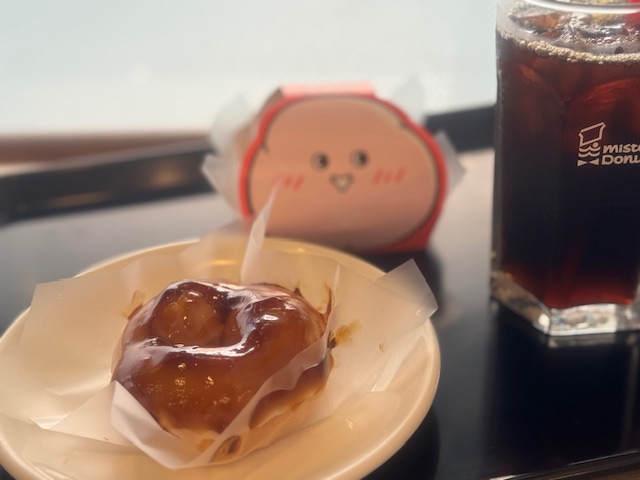 おさらのうえにのった、もっちゅりんのみたらしの写真。半透明のフィルムがしたにしかれています。ドーナツの表面にみたらしがかかっています。はいごにわ紙のケースのもっちゅりんと、グラスにハイッタアイスコーヒーがうつりこんでいます