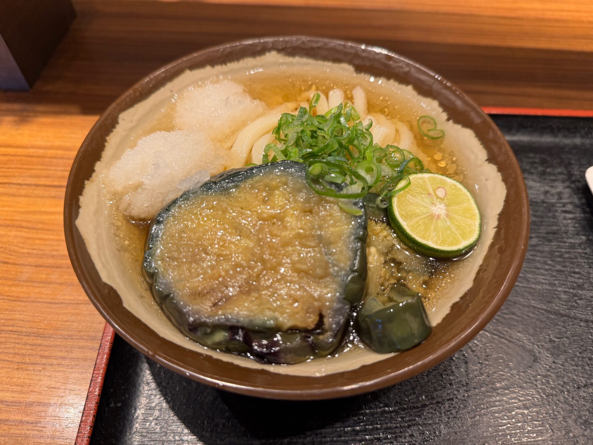 くろいおぼんにのった:なすみぞれひやかけうどん、なみサイズの写真。おおきなどんぶりに、うどん、だいこんおろし、ネギ、すだち、すあげされた、かなりおおきなわぎりのなすがのっています。ほぼうどんがみえないくらい、具だくさんです
