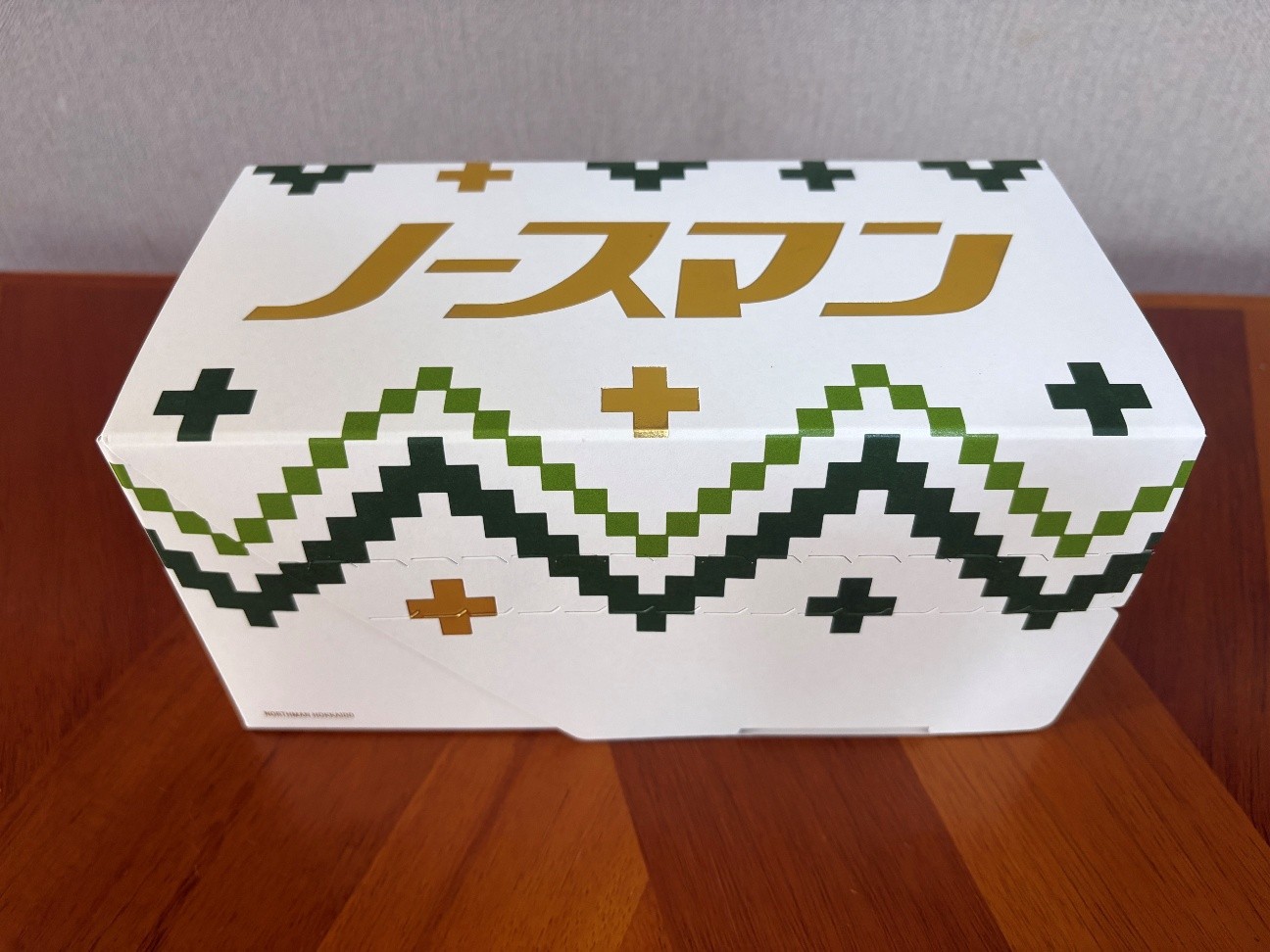 白い箱に金色の文字でノースマンと印字されたパッケージが、木目調のテーブルの上に置かれています。箱の側面には、緑色ときみどり色のドットで描かれた山のような模様が横一列に並んでおり、その上下には金色の十字のマークが配置されています。また、箱の左下隅には、小さくノースマンホッカイドウと英字で記載されています。箱全体は白を基調としており、シンプルで清潔感のあるデザインです。