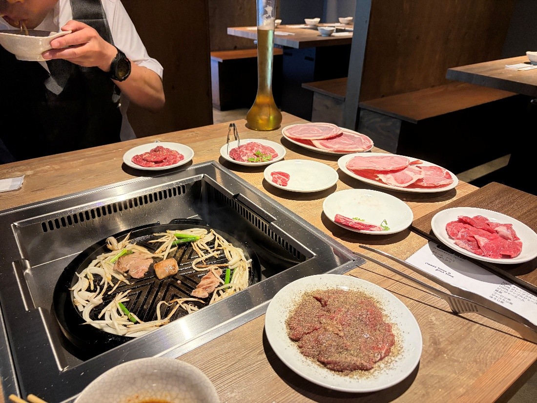 テーブルに並べられた羊肉や野菜、調理中の鉄板の様子です。中央には、中央が盛り上がった黒い鉄板が埋め込まれたテーブルがあり、鉄板の上では羊肉やもやし、ネギを焼いています。鉄板の周囲には、生の羊肉が盛られた白い皿が複数並んでいます。手前中央の皿には、黒胡椒がたっぷりとかかった赤身の羊肉が乗っています。中央奥には、背の高いグラスに入った飲み物が置かれています。テーブルの右側には、羊肉を焼くための金属製のトングが置かれています。